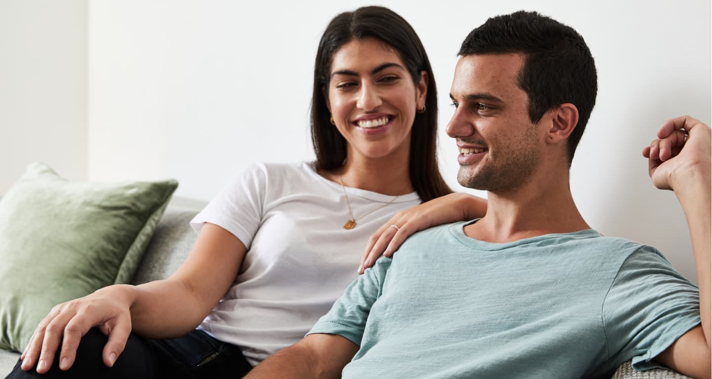Woman smiling at a man while they sit together on a couch. The man looks pleased about his full head of hair.